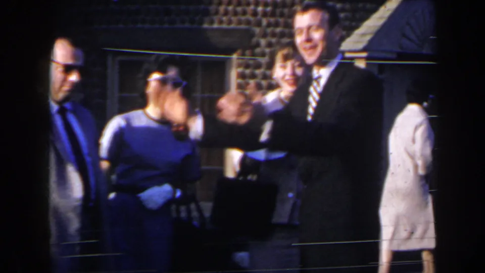 1963 Niagara Falls home-movie frame with a smiling man waving near women in dresses outside a brick facade.