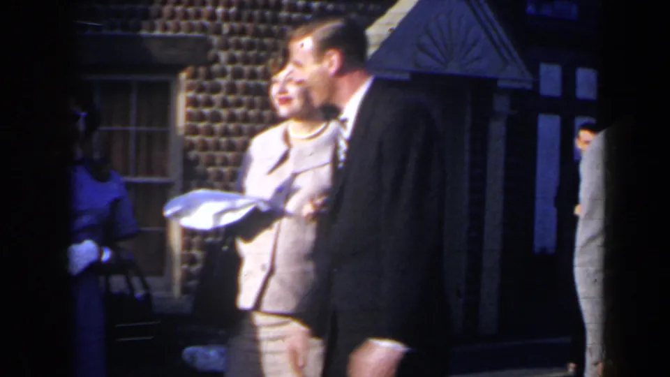 1963 Niagara Falls home-movie frame showing adults in dress clothes gathered by a brick building exterior.
