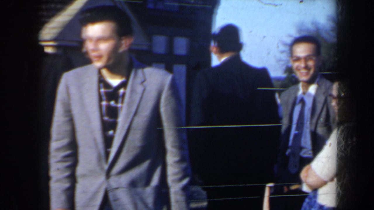 A young man in a gray suit stands outside near another man and a smiling woman.