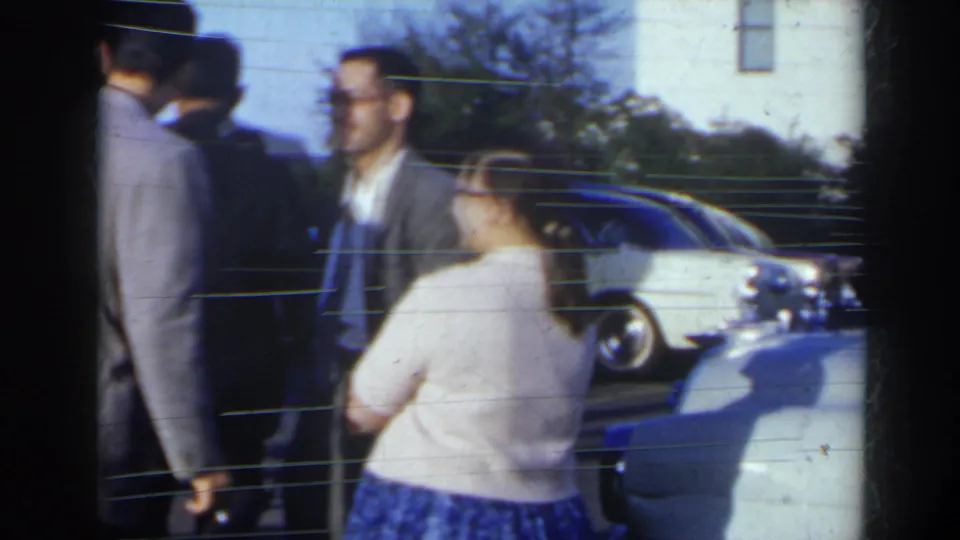 Outdoor 1963 home-movie frame with formally dressed adults standing near rounded parked cars in Niagra Falls, Canada.