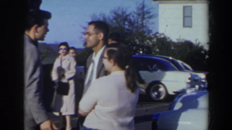 1963 Niagra Falls, Canada frame showing a small outdoor conversation among formally dressed adults beside parked cars.