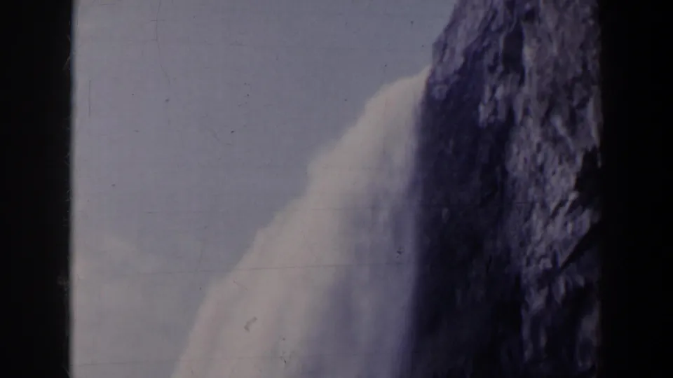 1963 visitor-shot 8mm frame of a waterfall dropping along a dark cliff, with grain and scratches from the film scan.