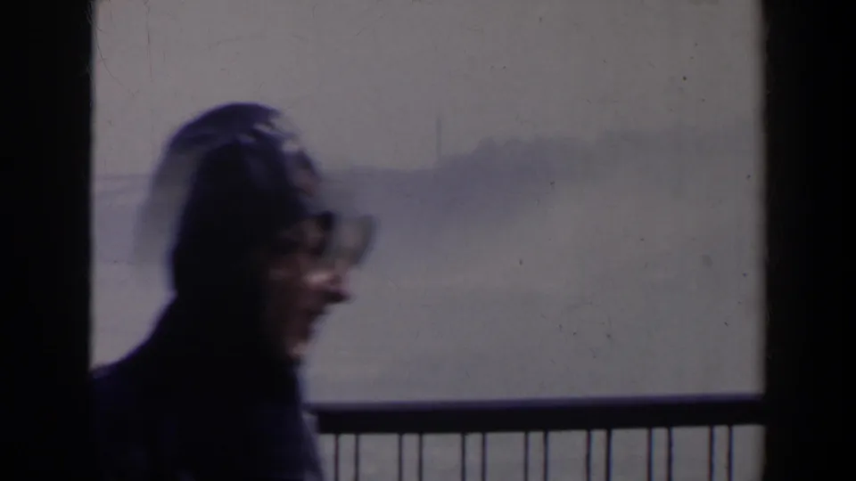 A hooded visitor wearing eyeglasses moves close to the camera at the Niagara Falls overlook, with railing and misty water behind.