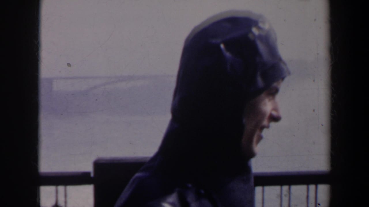 Person in a dark hooded coat stands near a railing with misty falls in the background.