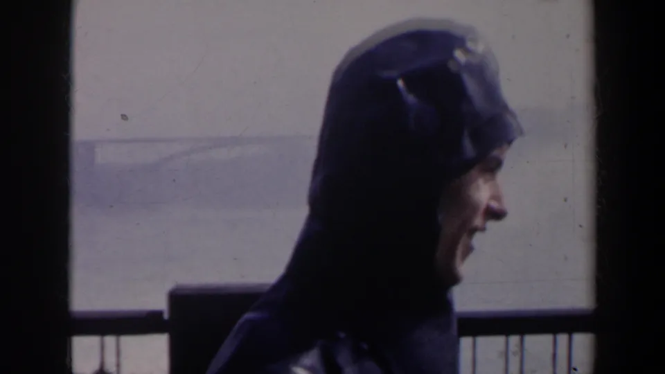 1963 Niagara Falls home-movie frame showing a person in a dark hooded coat in profile near a viewing railing and misty water.