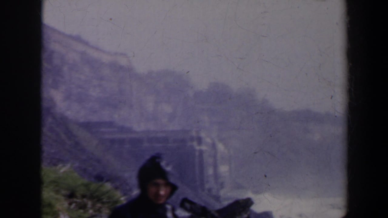 Two people near a misty waterfall overlook, one partly visible in a dark hooded coat.