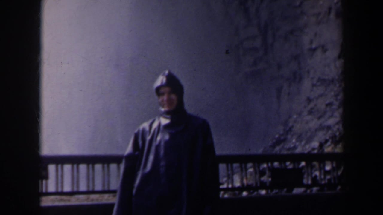 Person in a dark hooded raincoat stands by a railing with waterfall mist behind.