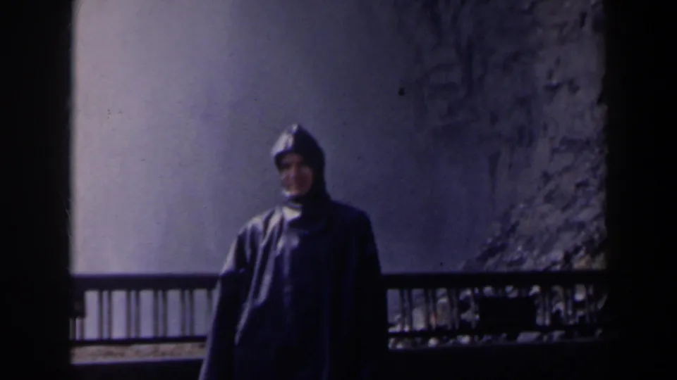 Person in a dark hooded raincoat stands at a Niagara Falls overlook with mist, cliff face and railing behind them.