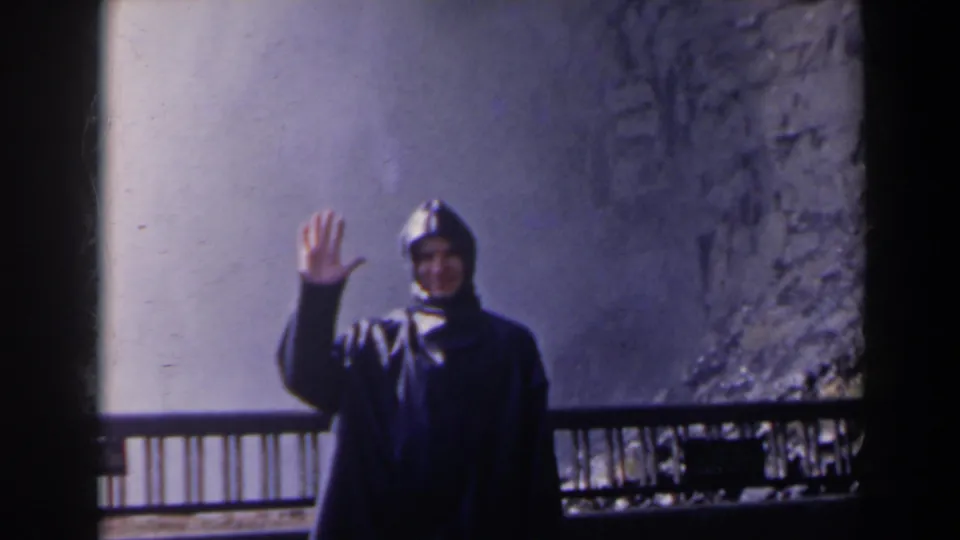 At Niagara Falls, Canada, a hooded visitor in a dark raincoat waves toward the 8mm camera beside the overlook railing.