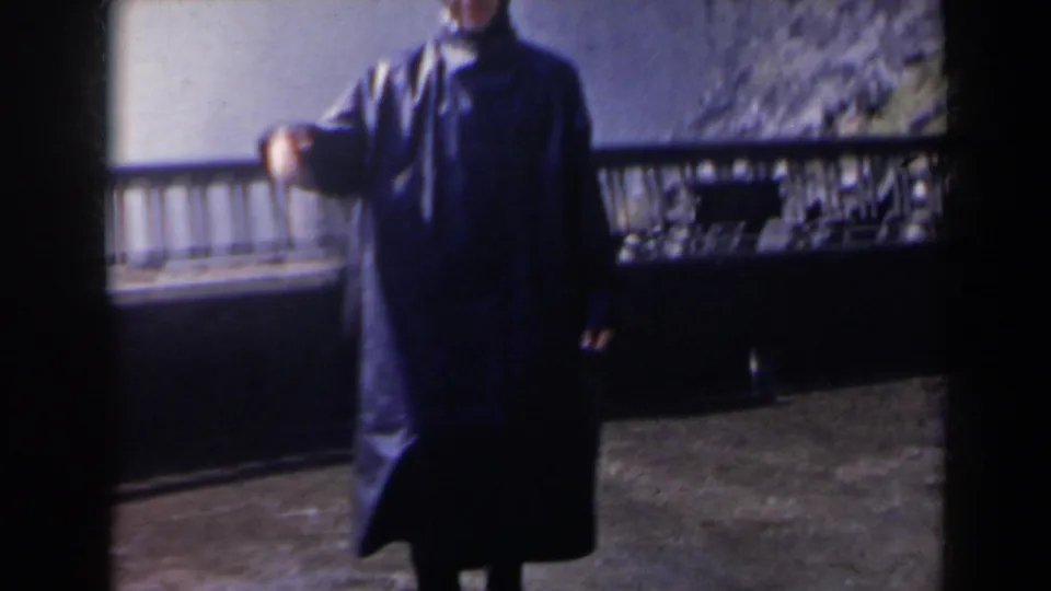 A hooded visitor in a long dark raincoat stands at the Niagara Falls overlook with mist, wet pavement and railing visible.
