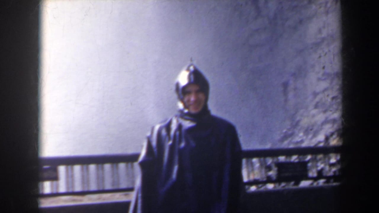 In 1963 Niagara Falls, Canada footage, a man in a dark hooded rain slicker faces the camera near a railing with pale waterfall spray behind him.