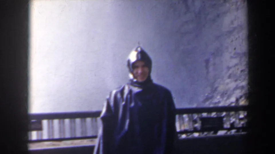 In 1963 Niagara Falls, Canada footage, a man in a dark hooded rain slicker faces the camera near a railing with pale waterfall spray behind him.