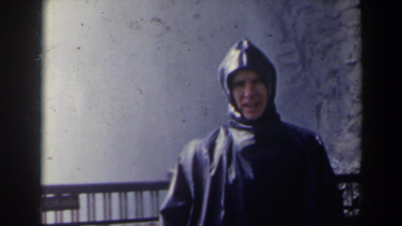 Closer view of a man in a hooded slicker near a railing with pale waterfall background.