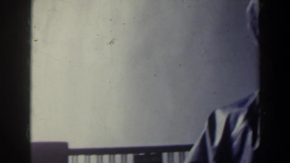The man moves partly out of frame in the Niagara Falls home movie, leaving a partial slicker, railing, pale wall, and dark film edge visible.