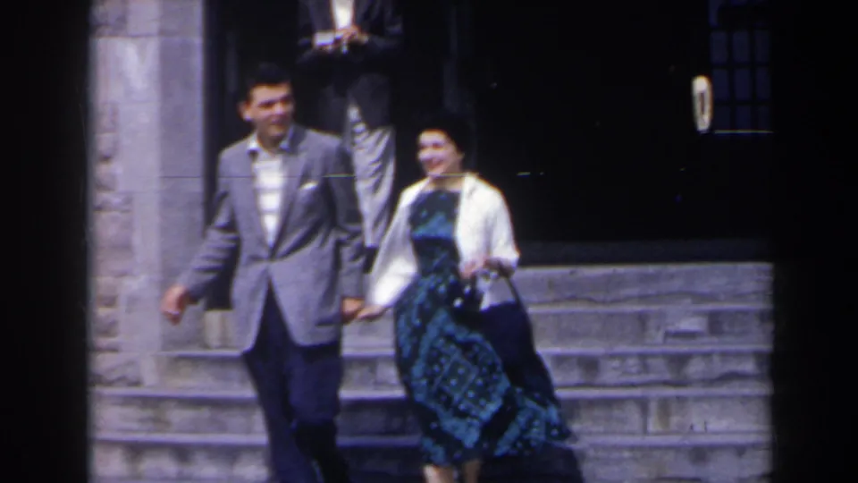 The couple descends the steps together, with the man in a light grey jacket and the woman in a dark patterned dress and white cardigan.
