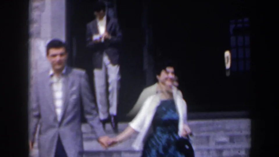 A closer 8mm frame shows the couple passing the camera while another adult stands near the dark building entrance behind them.