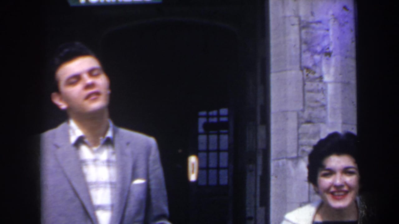 A young man in a gray suit stands smiling beside a woman near a stone entrance.