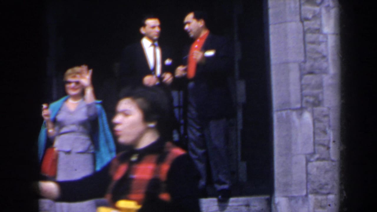1963 home-movie frame in Niagara Falls, Canada, showing adults on stone steps while a woman in gray and turquoise waves near the camera.