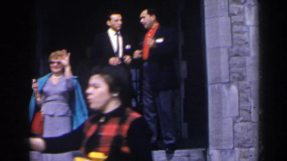 1963 home-movie frame in Niagara Falls, Canada, showing adults on stone steps while a woman in gray and turquoise waves near the camera.