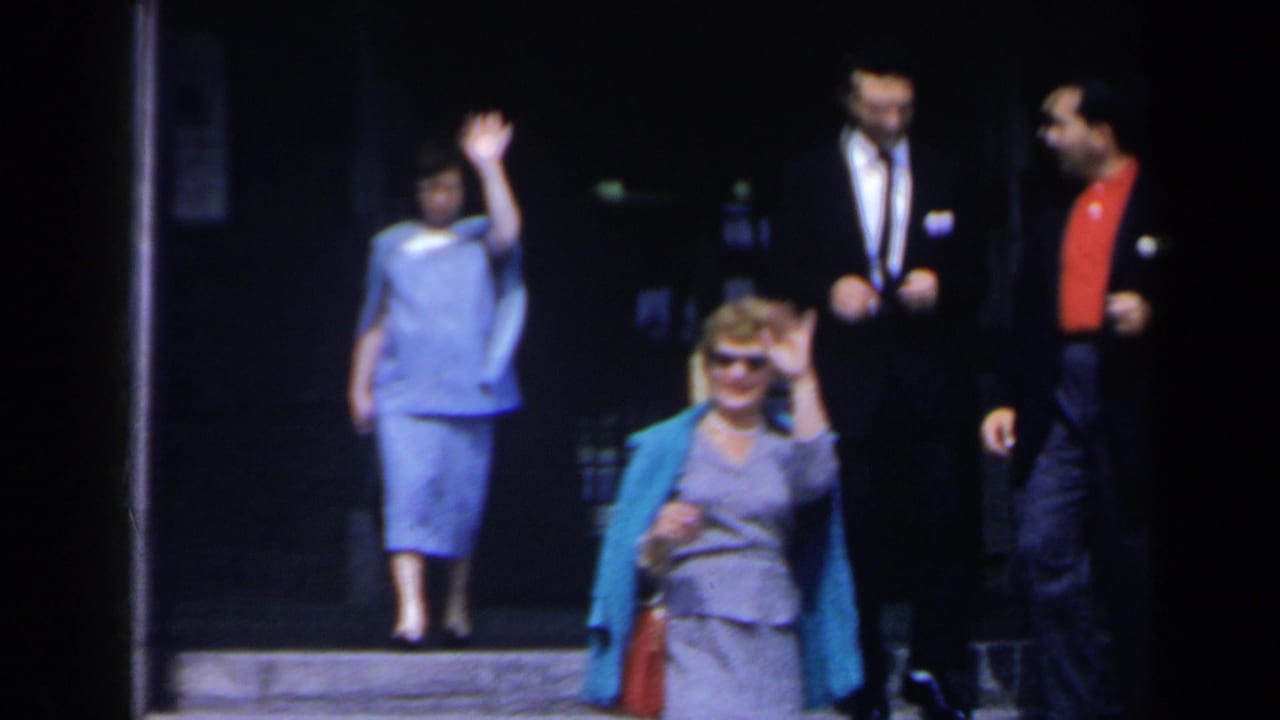 A woman in a light blue dress waves from a doorway as others stand and walk nearby.