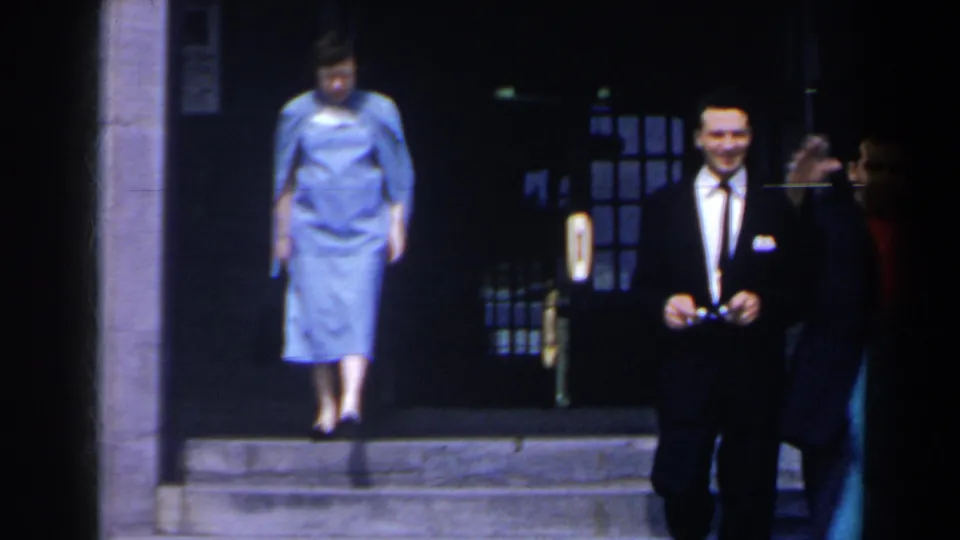 1963 Niagara Falls, Canada frame showing a woman in a light blue dress stepping down from a stone entrance near a suited man.