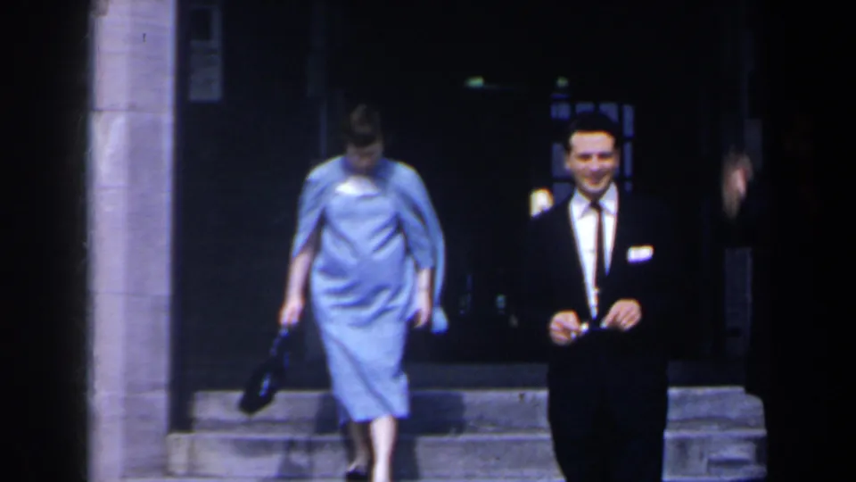 1963 Niagara Falls home-movie frame with a woman in a blue dress descending steps and a smiling suited man near the entrance.