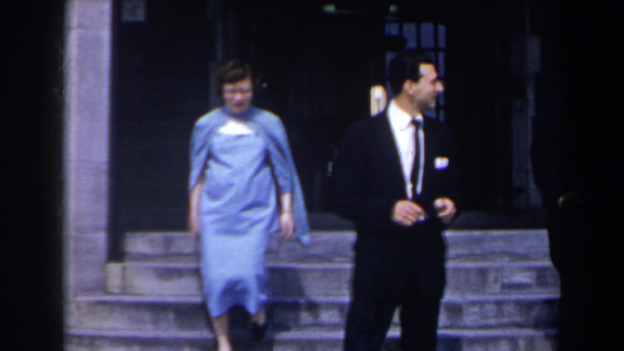 In 1963 Niagara Falls footage, a woman in a light blue dress exits behind a suited man near stone steps and a dark lobby entrance.