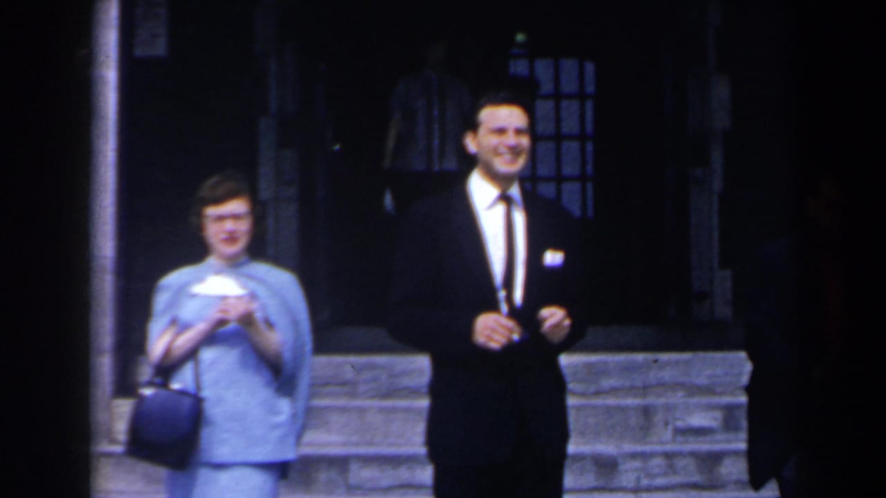 Smiling woman with handbag and suited man stand outside a dark lobby entrance.