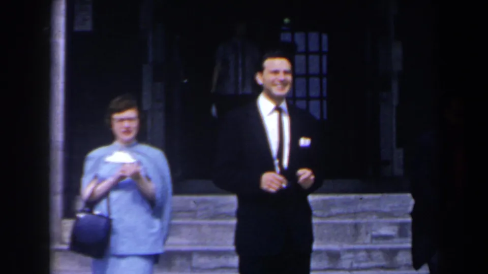 The woman in blue holds a handbag while the suited man smiles outside a Niagara Falls hotel entrance in 1963 home-movie footage.