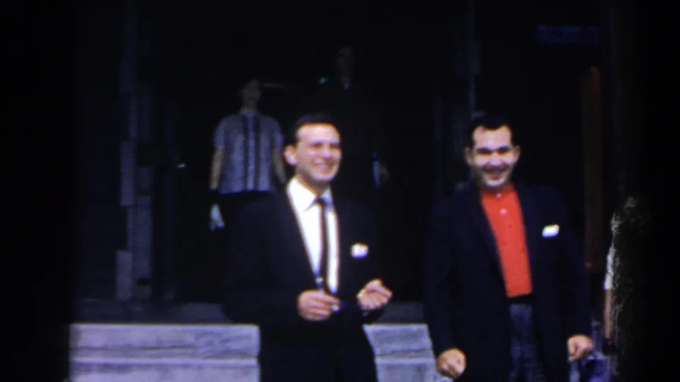 Two adult men, one in a dark suit and one in a dark jacket with red shirt, smile outside the lobby in 1963 Niagara Falls footage.
