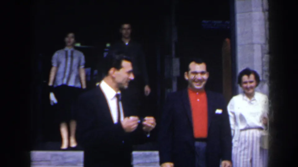 A suited man gestures beside another smiling man, with a woman in a light dress and lobby guests behind them in 1963 Niagara Falls footage.