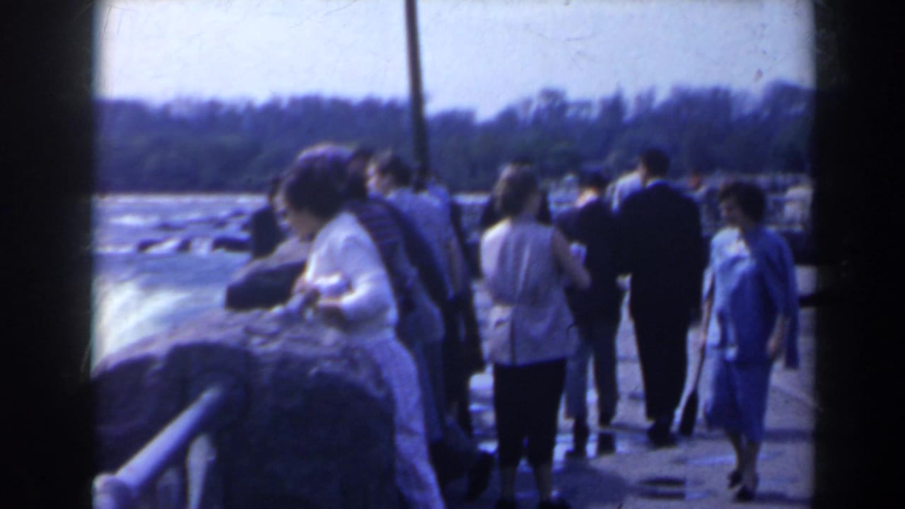 1963 home-movie view of tourists at a Niagara Falls overlook, with stone railing, rushing water, and distant shoreline visible.