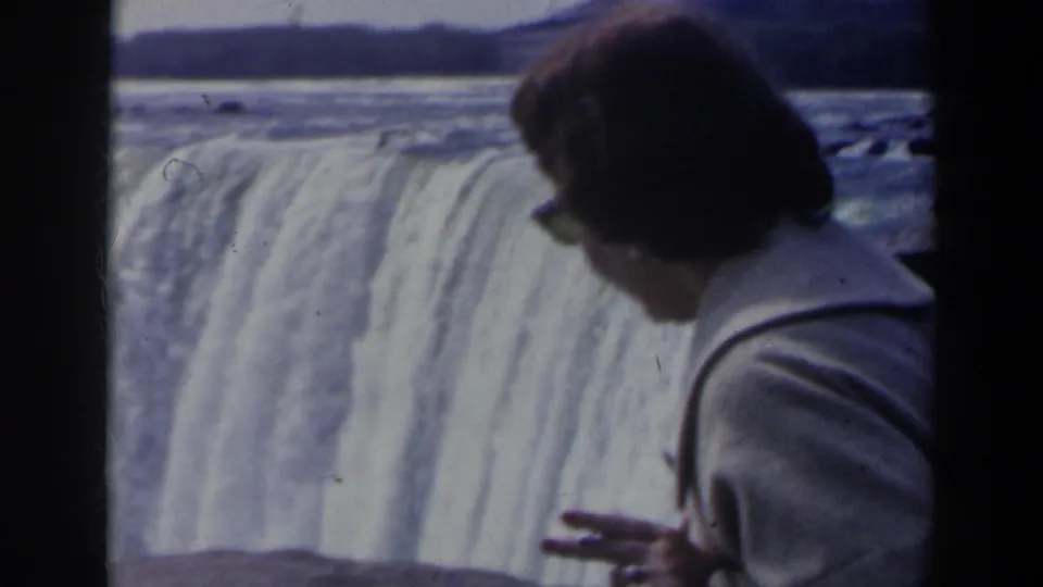 A woman in sunglasses appears in close foreground at the Niagara Falls overlook, with the curtain of water behind her.
