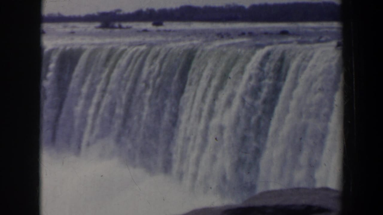 1963 8mm view of Niagara Falls from Canada, showing a broad waterfall crest, falling water, mist, and distant shoreline.