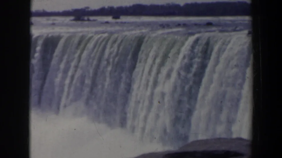 1963 8mm view of Niagara Falls from Canada, showing a broad waterfall crest, falling water, mist, and distant shoreline.