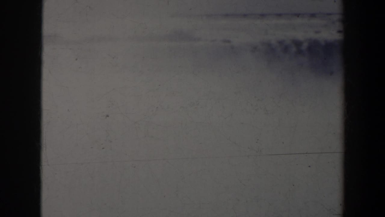 Dark scratched 8mm frame with pale gray exposure and black film borders.