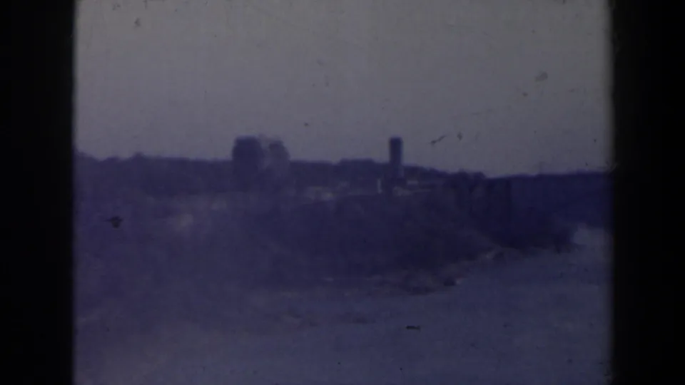 Blue-purple 8mm view of Niagara Falls, Canada, with mist, distant shoreline silhouettes, and low structures under a pale sky.