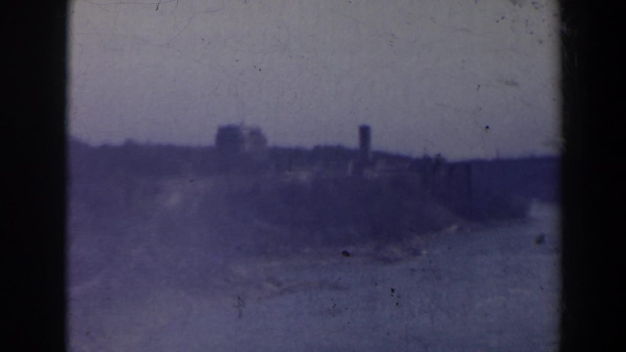 1963 8mm Niagara Falls area view with distant buildings, a tower-like form, haze, grain and dark frame-edge shadows.