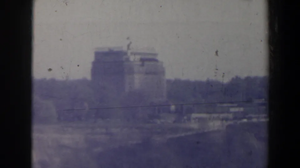 1963 home-movie frame of a distant multi-story structure near Niagara Falls with haze, grain and a dark horizontal element in front.