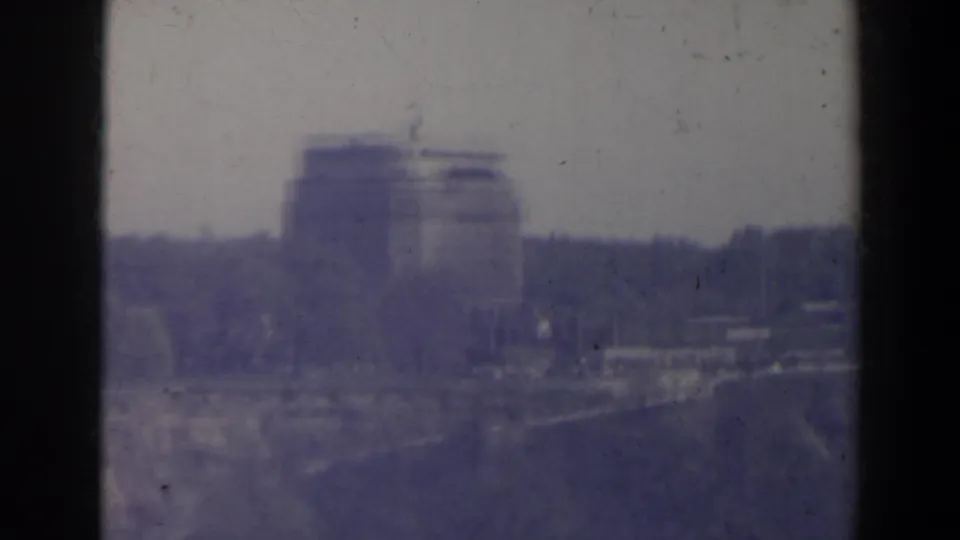 Hazy 1963 8mm Niagara Falls area view of a large distant building with dark trees and possible road or railing below.