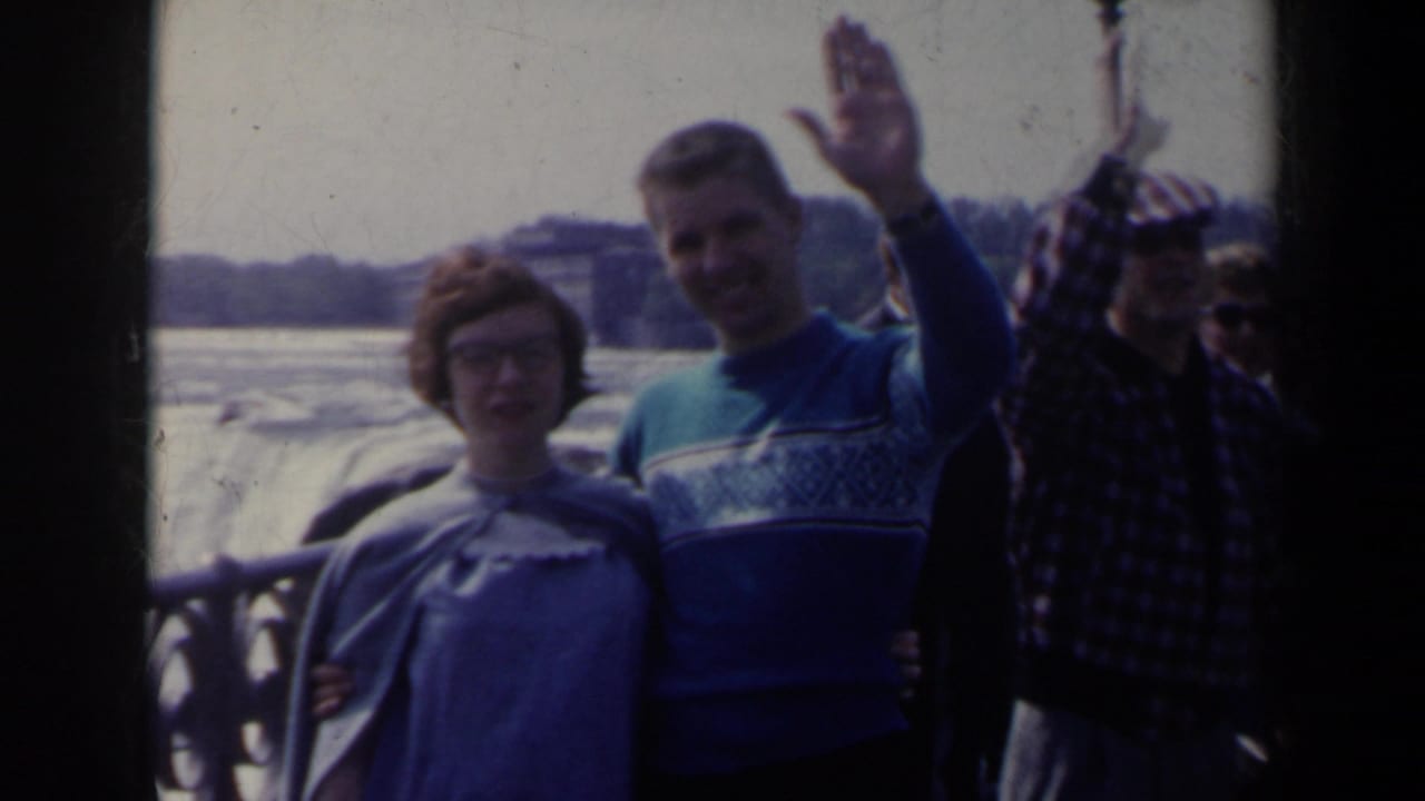 A young couple stands near a railing at Niagara Falls in Canada, with rushing water and other tourists visible behind them.