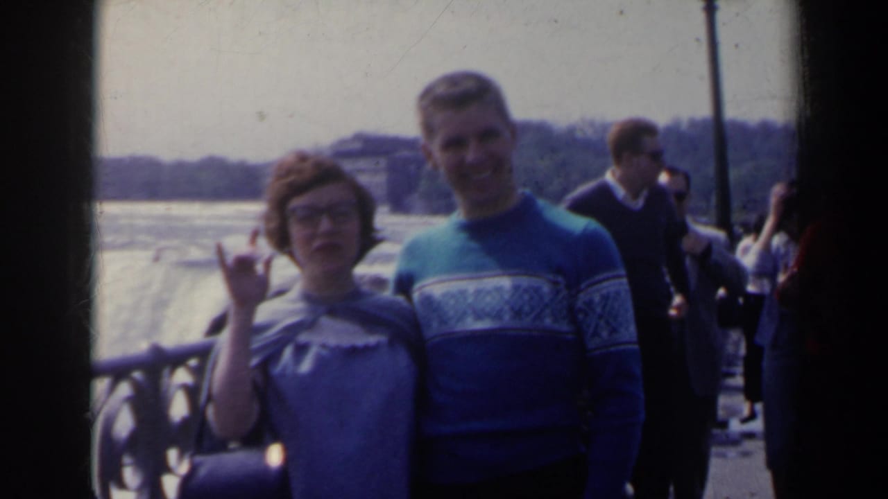 Couple beside a railing with falls, tourists, and lamp posts in the background.