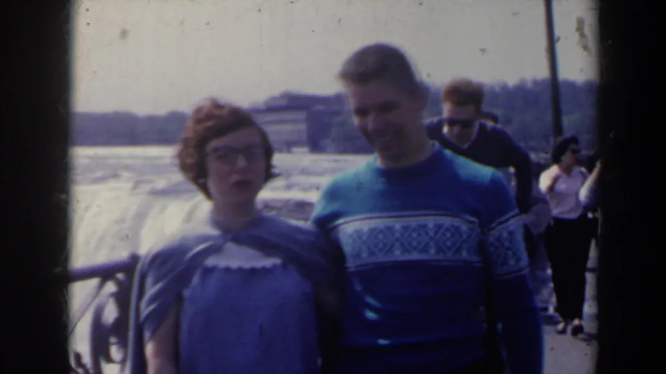 A young couple poses at a Niagara Falls overlook, with soft focus, railing details, and background tourists on the walkway.