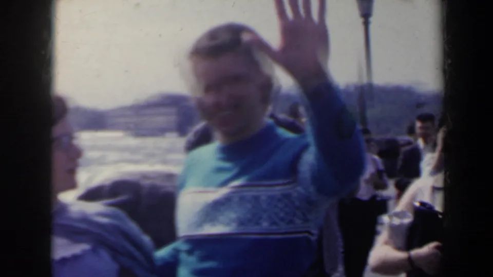 The man waves near the camera while the woman is partly out of frame, with Niagara Falls and tourists still visible behind them.