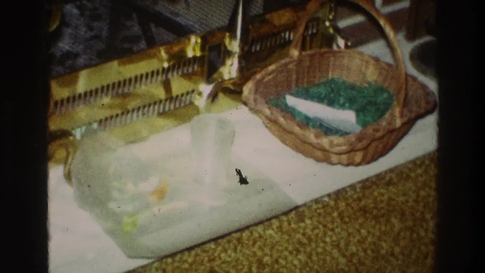 1969 Clarendon Hills home-movie frame showing a wicker Easter basket with green grass and a white egg-shaped object on a tray near brass fireplace tools.