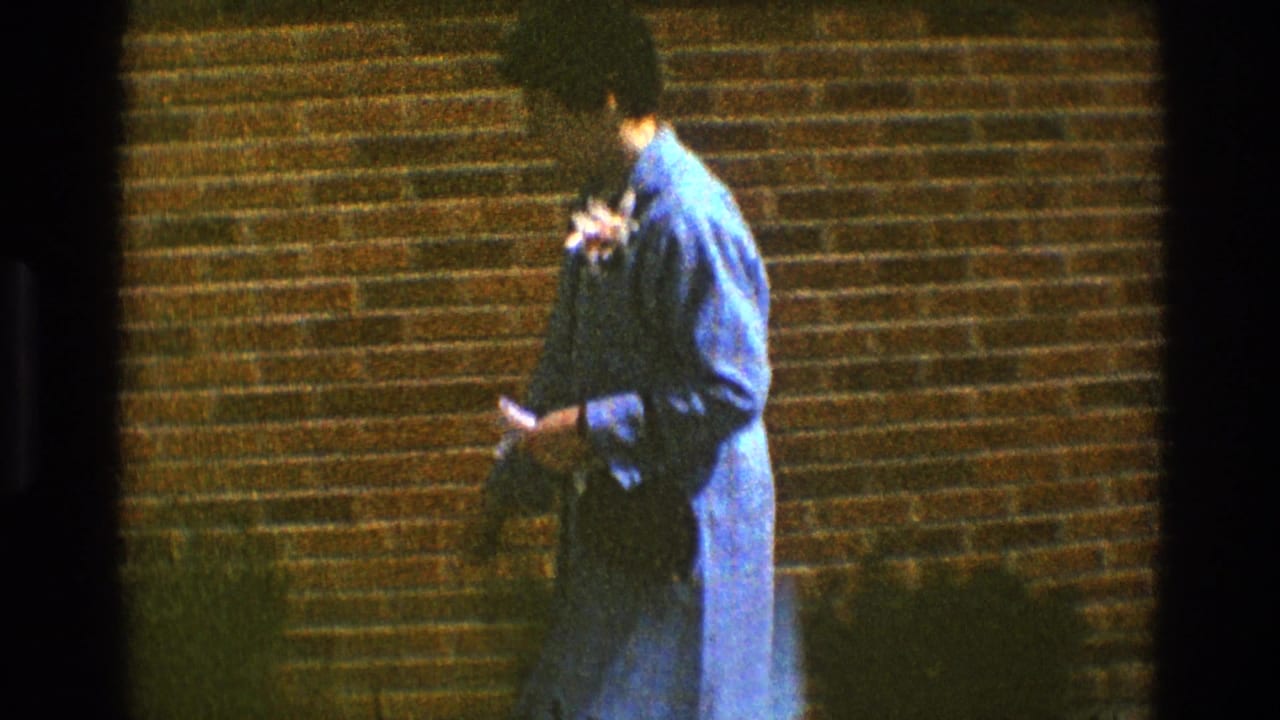 Woman in a blue dress walking beside a brick wall while holding dark fabric and flowers.