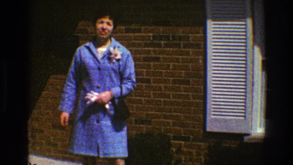 The woman faces the home-movie camera, holding fabric and flowers in front of a brick house with white shutters.