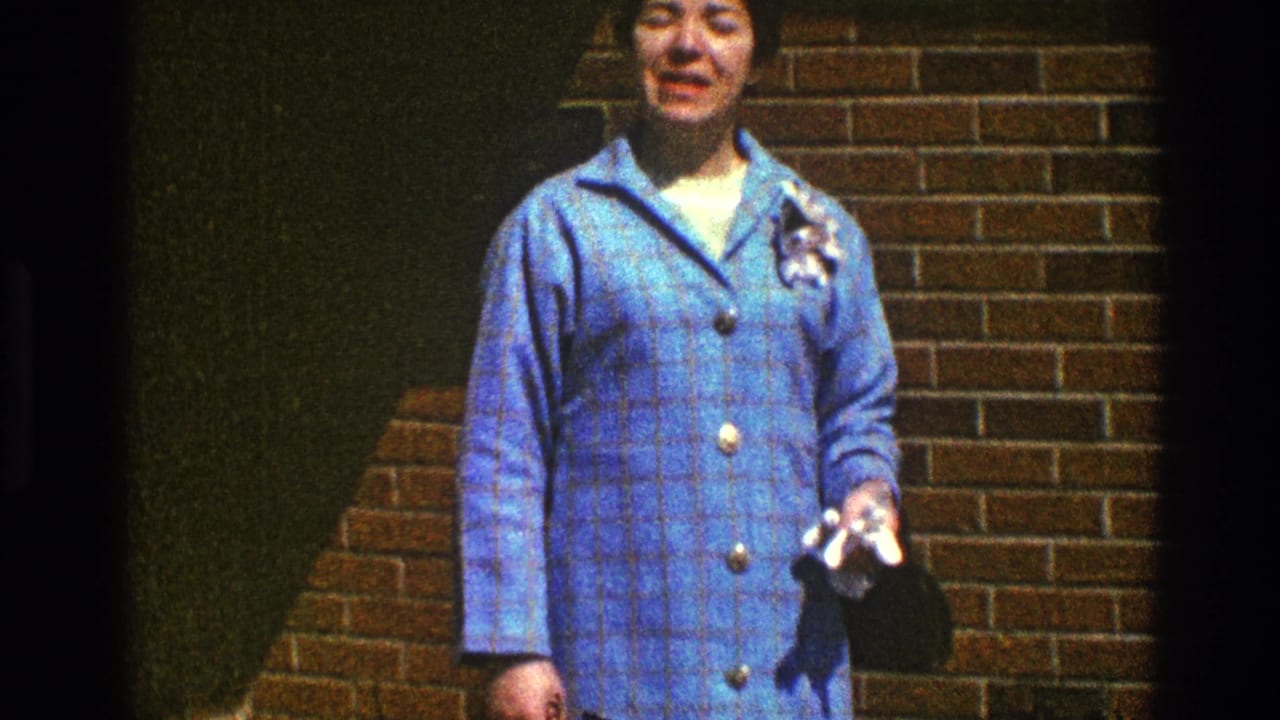 In Clarendon Hills, Illinois, a woman in a blue plaid coat with a flower pin faces the camera against a brick wall in 1969 home-movie footage.