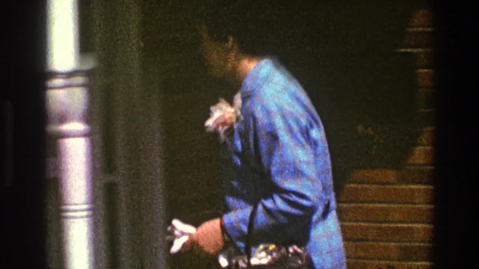 The woman turns in profile near a porch column, holding a small light object as she moves away from the brick wall.