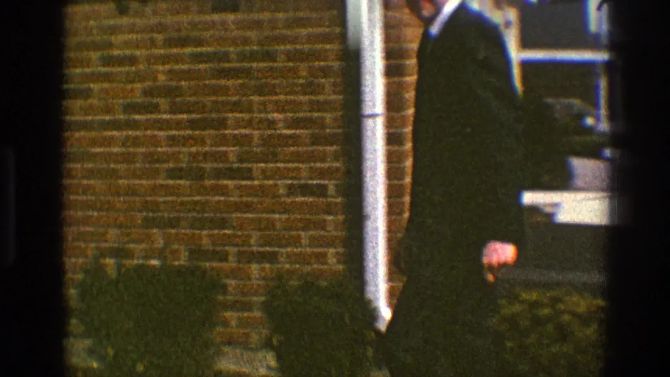 A man in a dark suit leaves the Clarendon Hills house exterior, passing the brick wall and white porch post in the 1969 home movie.